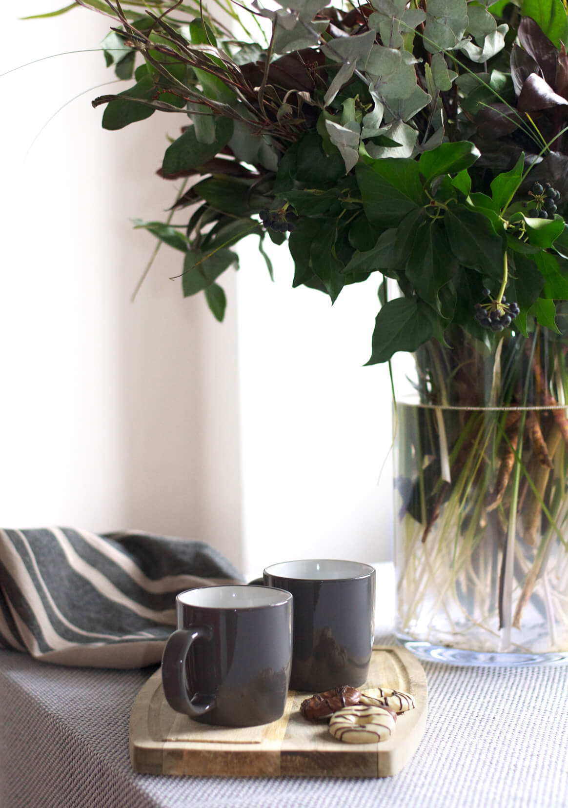 Close up of tea cups and biscuits with a bunch of greenery arranged in a vase in the background