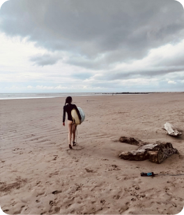 Sara walking on a beach with a surfboard